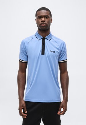 Bărbat stând, purtând un tricou polo albastru deschis cu o fundație din nasturi negri și logo-ul „BOSS” pe pieptul stâng.