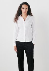 Camicia bianca con colletto classico, maniche lunghe e vestibilità su misura, abbinata a pantaloni neri con una texture liscia.