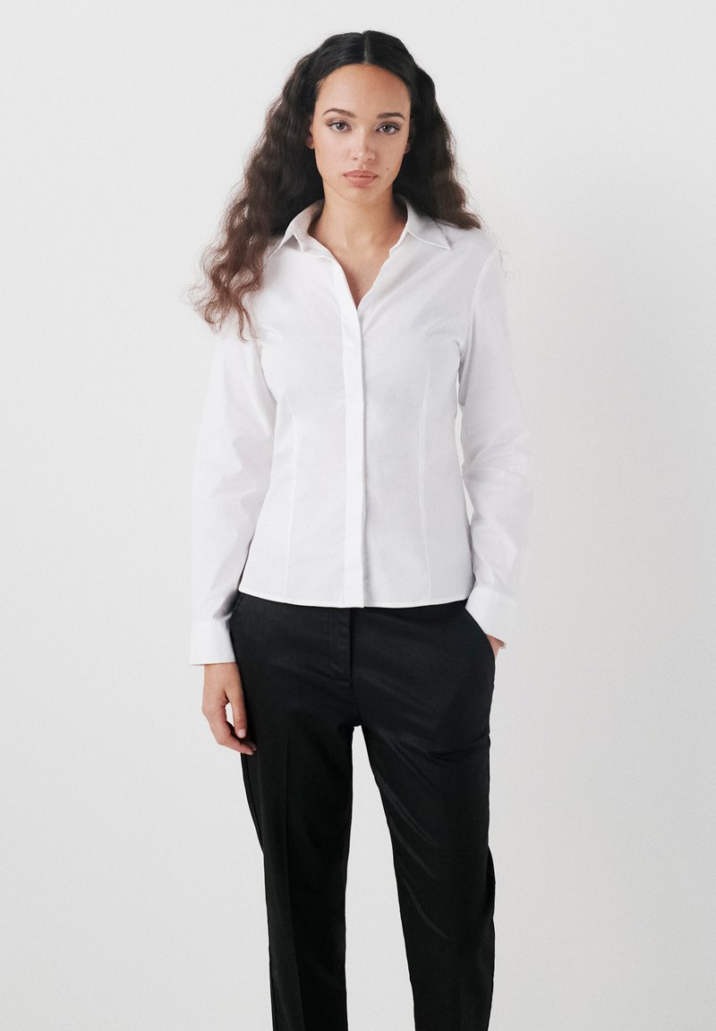 Camicia bianca con colletto classico, maniche lunghe e vestibilità su misura, abbinata a pantaloni neri con una texture liscia.