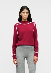 Pull bordeaux avec un col montant, des détails blancs en accent, et des manches longues ; associé à un pantalon gris ajusté. Texture en tricot doux.