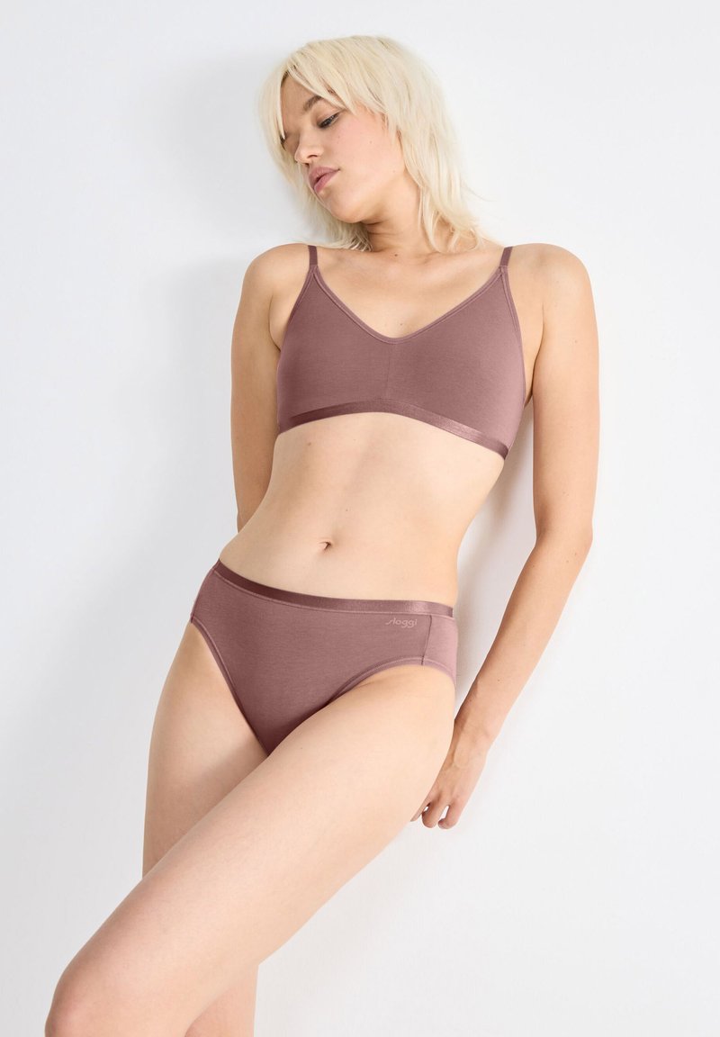 Donna con capelli biondi corti, che indossa un reggiseno malva e slip a vita alta, appoggiata a un muro bianco, con lo sguardo rivolto verso il basso.