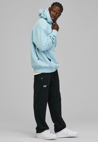 Lys blå oversized hettegenser med frontlomme, brukt sammen med svarte treningbukser med sidelommer og hvit grafisk tekst, kombinert med hvite sneakers.