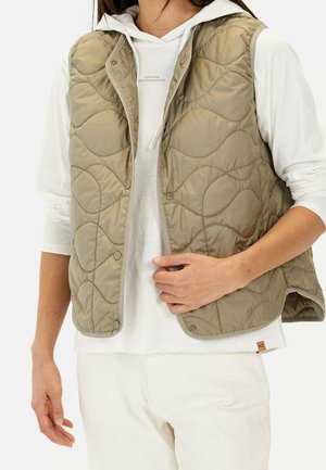 Personne portant un gilet matelassé olive par-dessus un sweat à capuche blanc et un pantalon blanc, ajustant l'avant du gilet d'une main.