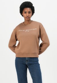 Hnědá mikina s žebrovanými manžetami a lemem, se znakem "TOMMY HILFIGER EST. 1985" v bílé barvě. V kombinaci s tmavými džíny.