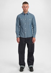 Camicia in denim azzurro chiaro a maniche lunghe con due tasche sul petto, chiusura a bottone e cuciture bianche, abbinata a pantaloni neri a gamba larga e scarpe nere.