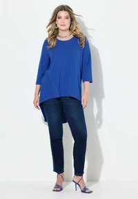 Top blu, morbido e ampio con maniche a 3/4 e orlo asimmetrico, abbinato a jeans in denim scuro con dettagli a bottoni e tacchi blu con cinturini.