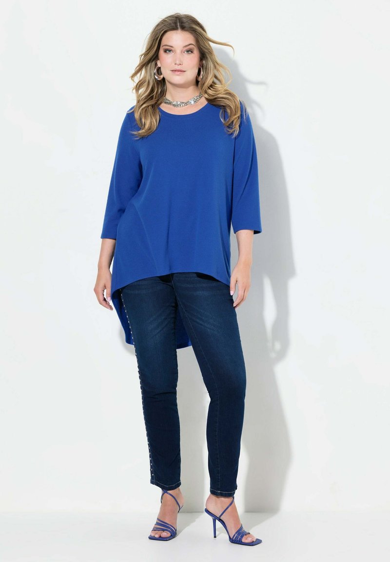 Top blu, morbido e ampio con maniche a 3/4 e orlo asimmetrico, abbinato a jeans in denim scuro con dettagli a bottoni e tacchi blu con cinturini.