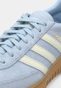 Ανοιχτό μπλε σατινέ αθλητικό παπούτσι με τρεις λευκές ρίγες, υφασμάτινο επάνω μέρος με υφή, σχέδιο με κορδόνια και καφέ λαστιχένια σόλα. Φέρει χρυσά γράμματα με την επιγραφή "SPEZIAL".