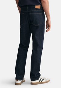 Jeans in denim blu scuro con taglio dritto, cuciture visibili e un patch con il logo GANT sulla vita posteriore, abbinati a sneaker bianche e nere.