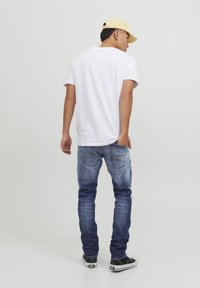 T-shirt en coton blanc, jeans en denim bleu avec des détails délavés, et baskets noires. La personne porte une casquette jaune clair, de dos.