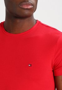 Mann in einem einfarbigen roten T-Shirt mit kurzen Ärmeln und einem kleinen rechteckigen Logo auf der linken Brust vor einem weißen Hintergrund.