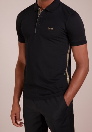 Mężczyzna ubrany w czarną krótkorękawową koszulkę polo Hugo Boss z złotym logo na piersi i złotym paskiem po boku, czarne spodnie, stojący z jedną ręką w kieszeni.