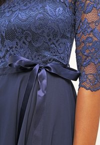 Une robe bleu marine avec un corsage en dentelle à motifs floraux, une ceinture en satin nouée à la taille et un tissu fluide.