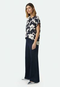 Luftige Bluse mit kurzen Ärmeln in blau-creamfarbenem abstrakten Muster, kombiniert mit dunkelblauen weit geschnittenen Jeans. Das Modell trägt silberne Schuhe.