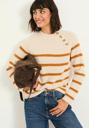 Cremfarbener Strickpullover mit rost-orangenen Streifen, rundem Halsausschnitt und seitlichen Knopfdetails. Kombiniert mit blauen Jeans und einer braunen Tasche.