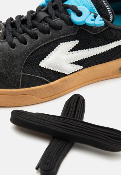 Μαύρο αθλητικό παπούτσι με λευκό λογότυπο βέλους, καφέ σόλα, μαύρα κορδόνια και μπλε κλειδώματα κορδονιών, συνοδευόμενο από ένα επιπλέον σετ μαύρων κορδονιών σε λευκό φόντο.