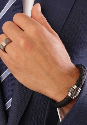 Bracelet en cuir tressé noir avec accent en argent, porté sur une main tenant une veste de costume, avec un fermoir en métal détaillé.