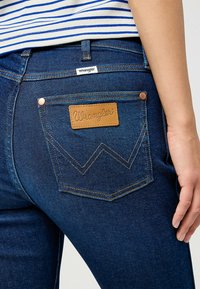 Blå denimjeans med en brun läderpatch från Wrangler på bakfickan, med guldtrådsställning och en minimalistisk design.