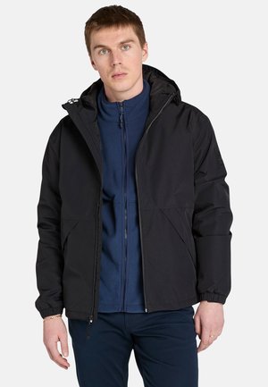 Un giovane con i capelli corti indossa una giacca nera con cappuccio sopra un maglione in pile blu, posando davanti a uno sfondo bianco semplice.