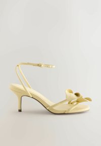 Sandale beige à petit talon avec bride cheville et boucle, avec un détail fleur décoratif sur la bride des orteils.