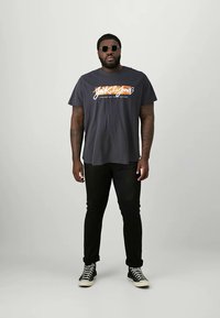 Schwarzes T-Shirt mit einem grafischen Logo in Orange und Weiß; kombiniert mit schwarzen Jeans und schwarzen High-Top-Sneakern. Die Person trägt Sonnenbrillen.