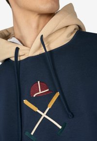 Felpa blu navy con cappuccio beige, dotata di ricami di un cappellino da baseball e di mazze incrociate in bordeaux, giallo e verde. Texture morbida.