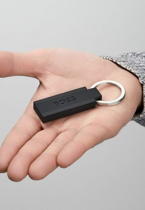 Musta suorakulmainen avaimenperä, joka on valmistettu teksturoidusta kumista ja jossa on hopeinen metallirengas. Sanat "BOSS" on kohokuvioitu pinnalle.