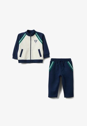 Completo tuta blu navy con giacca con zip e pantaloni. La giacca presenta una parte anteriore color crema, strisce verdi e bianche, e una grafica di Topolino.