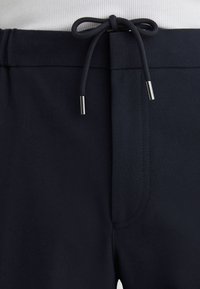 Black pants waistband with a tied black drawstring featuring metal aglets and a white ribbed shirt tucked in above.