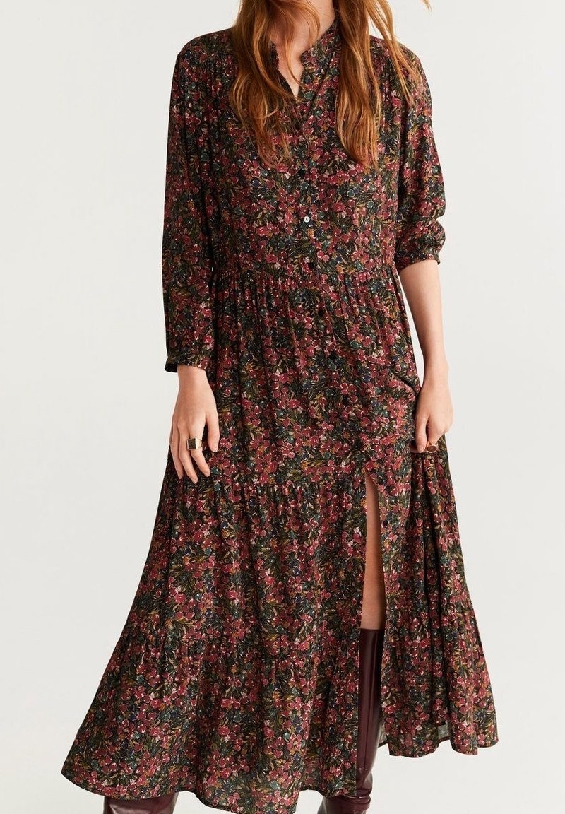 Robe longue fleurie aux tons rouge foncé, vert et marron, avec des boutons et une fente sur le côté, portée avec des bottes marron montant au genou.