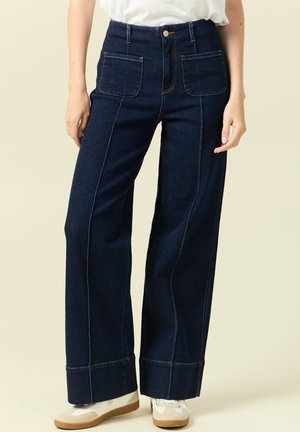 Kobieta w ciemnoniebieskich, szerokich dżinsach z wysokim stanem i przednimi kieszeniami naszywanymi oraz białych trampkach na beżowym tle.