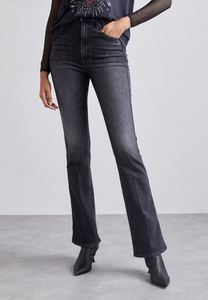 Personne portant un jean bootcut noir délavé, des bottines noires à bouts pointus et un top graphique noir avec manches longues en résille, debout sur un sol gris.