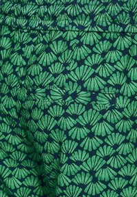 Zelená látka s opakujícím se vzorem listů v různých odstínech, umístěná na tmavě modrém pozadí. Obsahuje malý detail kapsy.