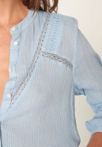 Lichtblauwe overhemd met knopen in een gestructureerde stof, met kant details langs de halslijn en een ruchesaccent aan de voorkant.