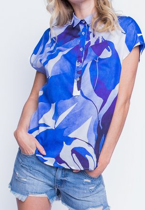 Nainen, jolla on yllään sinivalkoinen abstrakti kuvioitu lyhythihainen pusero, joka on hieman sullottu kuluneisiin farkkushortseihin, ja kädet taskuissa.