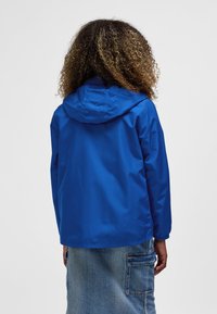 Blå, vandtæt jakke med hætte, der har en glat tekstur og elastiske manchetter. Kombiner med et denimskørt for et lagdelt look.