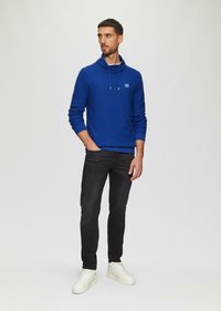 Modrá pletená mikina s kapucňou a šnúrkou, s logom na hrudi; v kombinácii s čiernymi slim-fit džínsami a bielymi teniskami.