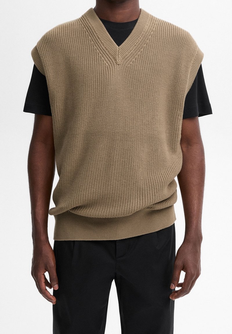 Beige knitted sleeveless sweater with a V-neck and textured fabric. Worn over a black T-shirt, paired with black trousers.