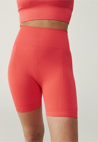 Kratší aktivní šortky ve vysokém pasu v korálové barvě s jemným texturou a přiléhavým střihem. Obsahují hladký pas a délku po střední stehna.