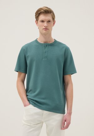 Joven con cabello castaño claro que lleva una camisa henley de manga corta color verde azulado y pantalones beige claros, de pie contra un fondo liso.