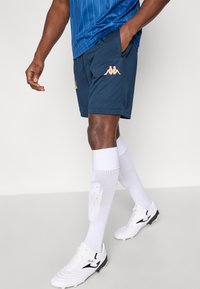 Tengerészkék sportnadrág oldalzsebekkel és rózsaszín logóval, fehér zoknival és fekete-fehér stoplis cipővel párosítva. Kék csíkos sporting.