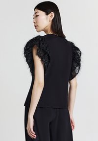 Top nero con maniche in tessuto trasparente arricciato e testurizzato. Il corpo è liscio e aderente, con orlo dritto e apertura sottile sul collo.