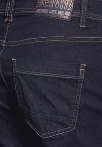 Vaqueros de mezclilla azul marino con un bolsillo trasero visible, costuras amarillas en contraste y una etiqueta negra con la palabra "DENIM" impresa en ella.