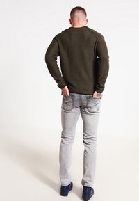 Pull tricoté vert olive à manches longues, assorti à un jean gris clair et des chaussures bleu foncé. Vue de dos, montrant les coutures détaillées.