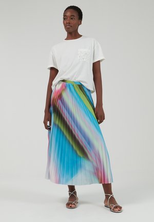 T-shirt blanc avec une poche à motif associé à une longue jupe plissée multicolore dans des teintes de bleu, vert et rose. Sandales à lanières.
