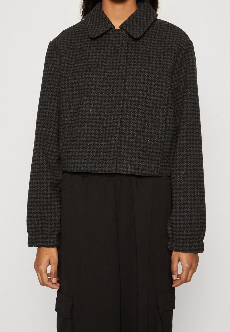 Personne portant une veste courte à manches longues motif pied-de-poule noir et gris sur une robe noire, bras détendus le long du corps.