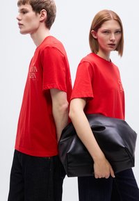 Κόκκινα βαμβακερά T-shirt με λογότυπο στο στήθος, συνδυασμένα με σκούρο τζιν. Μια μαύρη δερμάτινη τσάντα κρατιέται από ένα μοντέλο, με απαλή, ζαρωμένη υφή.