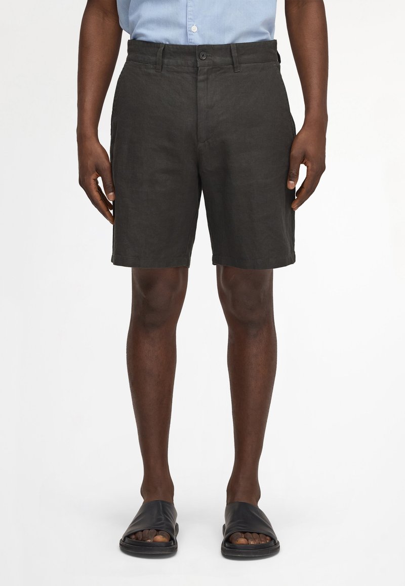 Mann trägt schwarze kniehohe Shorts, hellblaues Hemd mit Knöpfen, in die Hose gesteckt, und schwarze offene Sandalen, steht vor weißem Hintergrund.