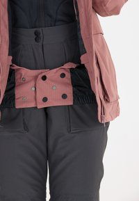 Rosafarbene Jacke mit schwarzer Innenauskleidung, ausgestattet mit Druckknöpfen an der Taille und einem strukturierten Design. Dunkelgraue Hose mit Taschen.