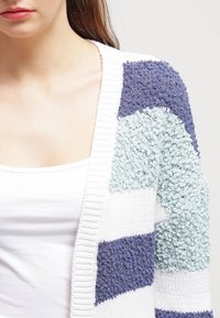 Le cardigan en tricot présente des rayures en zigzag texturées bleu, vert et blanc, avec un col côtelé et un tissu doux pour le confort.